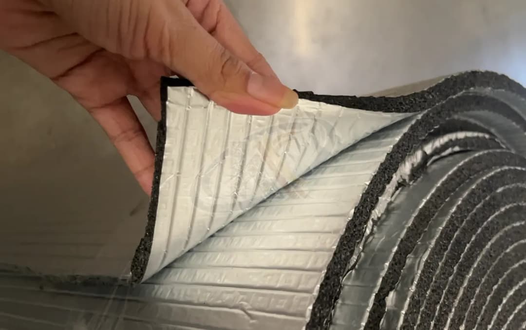 Foto realistis gulungan XLPE foam berlapis aluminium foil yang sebagian dibuka di atas meja kerja. Tampilkan potongan samping material untuk memperlihatkan ketebalan lapisan foam dan permukaan foil. Gunakan foto produk Karpinux sebagai referensi bentuk se